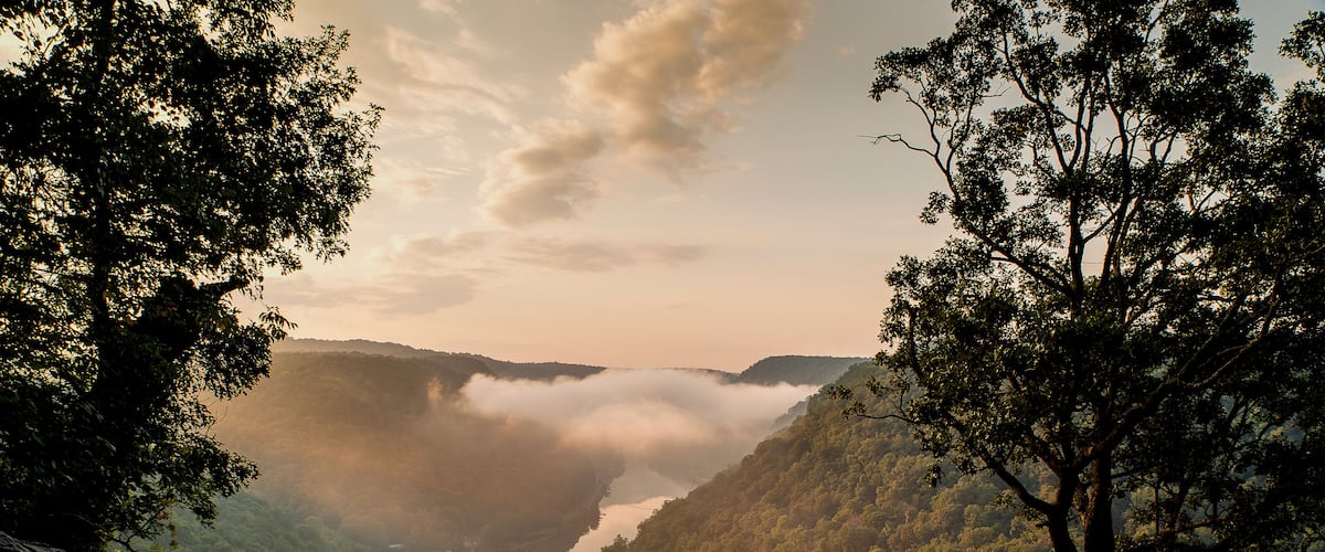 Un mirador pintoresco con un muro de piedra, un banco de madera y vistas a un río con un puente y colinas cubiertas de niebla.