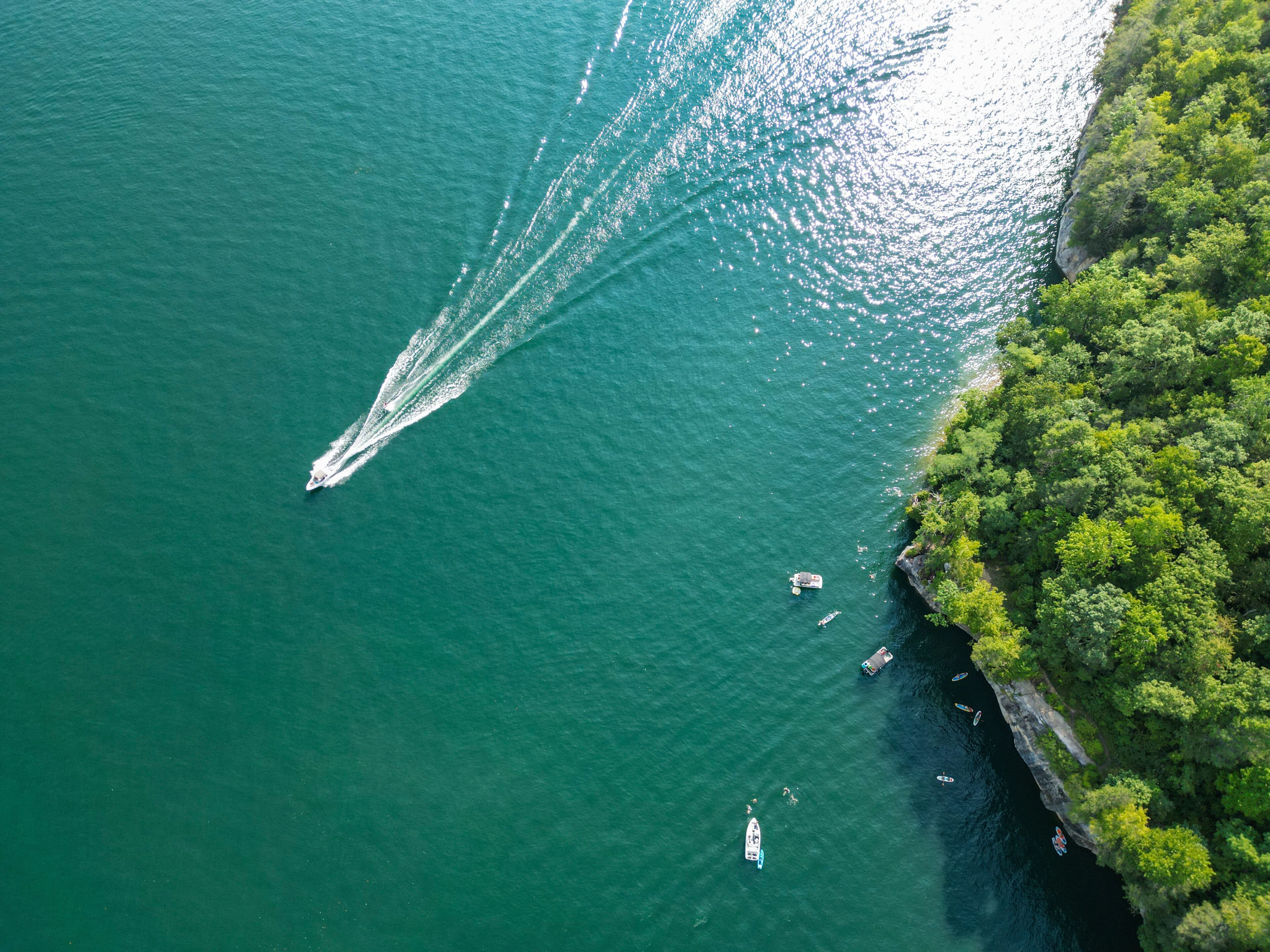 A boat creating a wake in a greenish-blue body of water, surrounded by dense green foliage and a rocky shoreline.