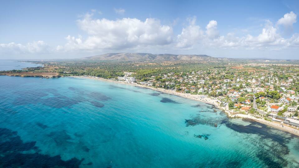 A coastal town with a beach, clear blue waters, and mountains in the background.
