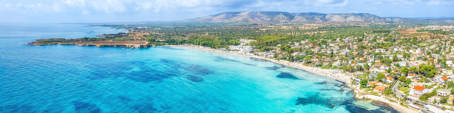 Un pueblo costero con mar azul cristalino, una playa de arena y montañas al fondo.