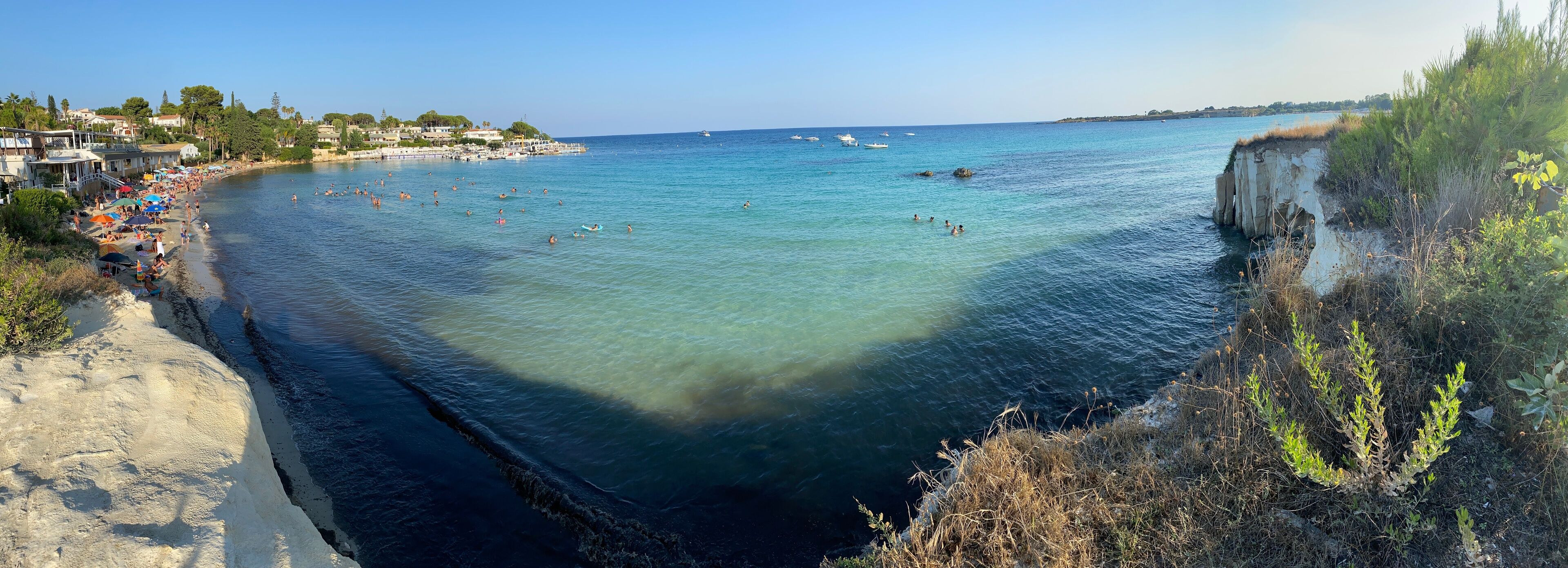 A coastal beach with clear waters, a rocky cliff, and a town in the background.