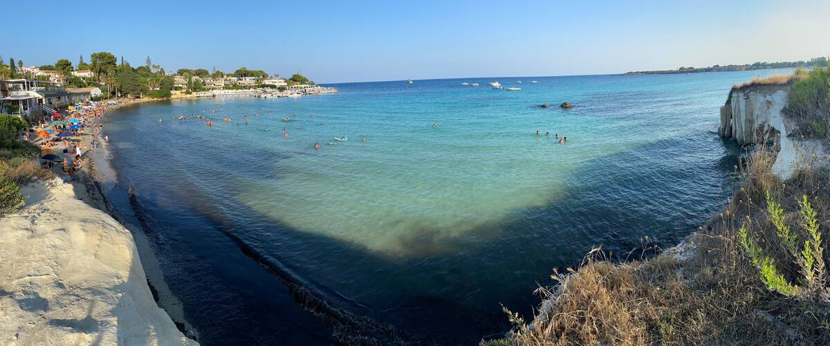 A coastal beach with clear waters, a rocky cliff, and a town in the background.