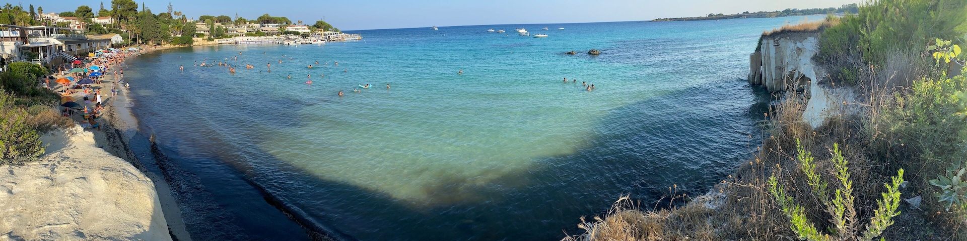 A coastal beach with clear waters, a rocky cliff, and a town in the background.