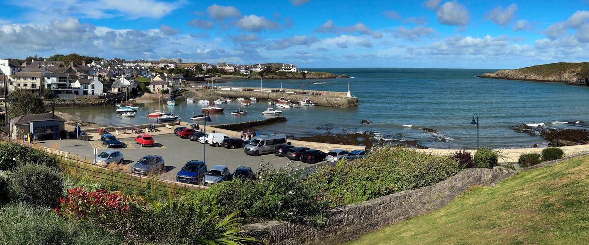 A coastal town with a marina, parked cars, and a grassy hill.