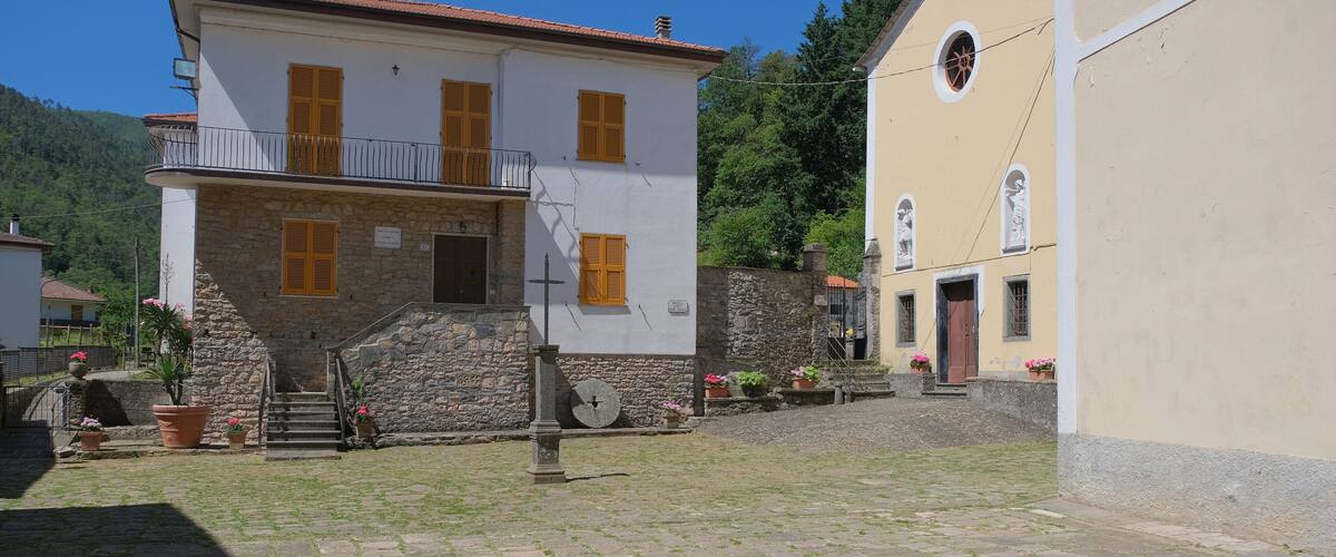 A stone building with a white facade and yellow shutters, surrounded by a cobblestone courtyard and lush greenery.