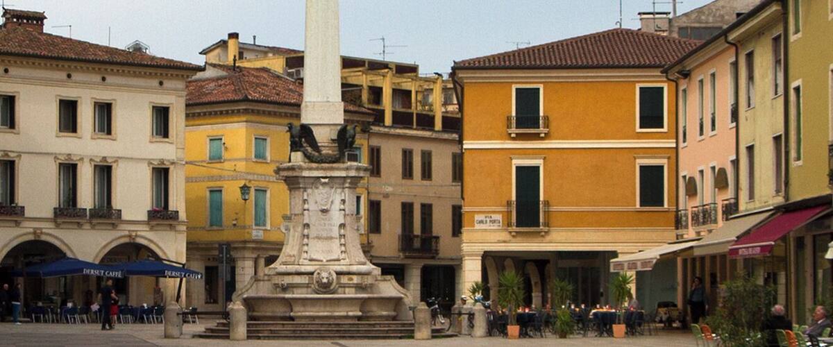 A narrow street with historic buildings, outdoor cafes, and a central monument.