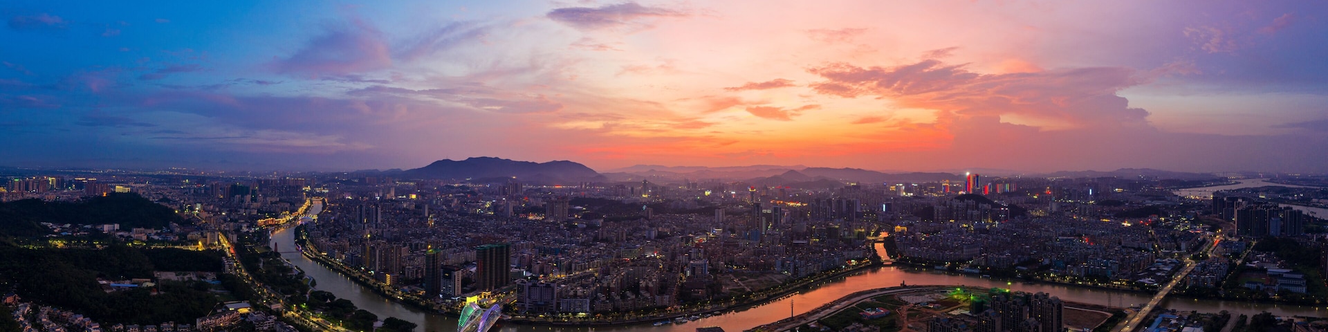 Paisagem urbana ao entardecer, com um rio serpenteando pela cidade, cercada por edifícios altos e uma ponte.