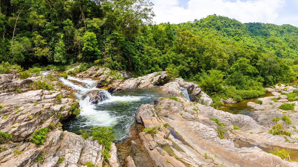 A river flowing over rocky terrain with lush green vegetation and a forested hill in the background.
