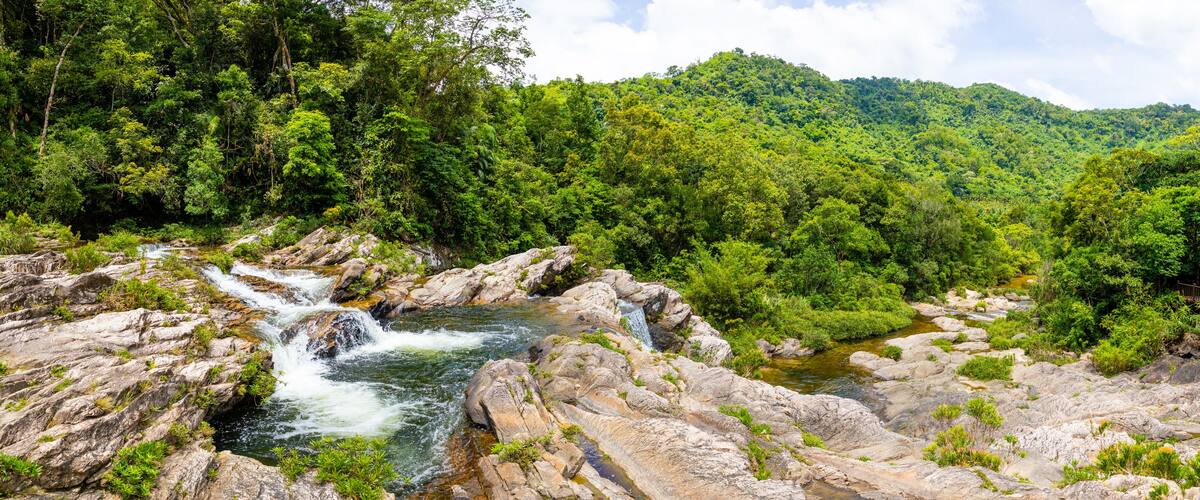 A river flowing over rocky terrain with lush green vegetation and a forested hill in the background.