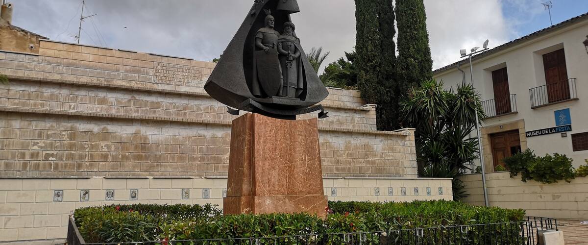 A statue of a woman holding a child, surrounded by a metal fence and greenery, with a building labeled 'Museu de la Festa' in the background.