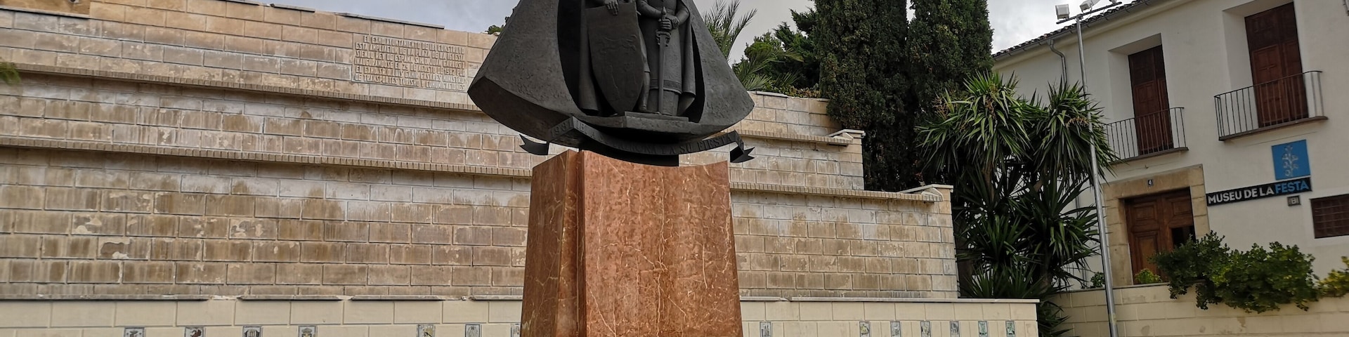 A statue of a woman holding a child, surrounded by a metal fence and greenery, with a building labeled 'Museu de la Festa' in the background.