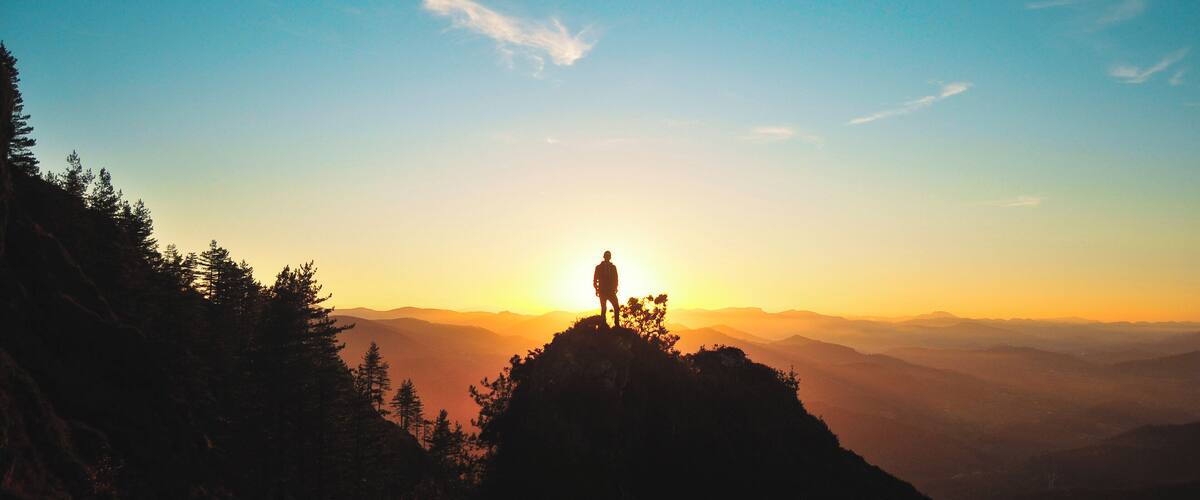 A person standing on a mountain peak with a sunset in the background.