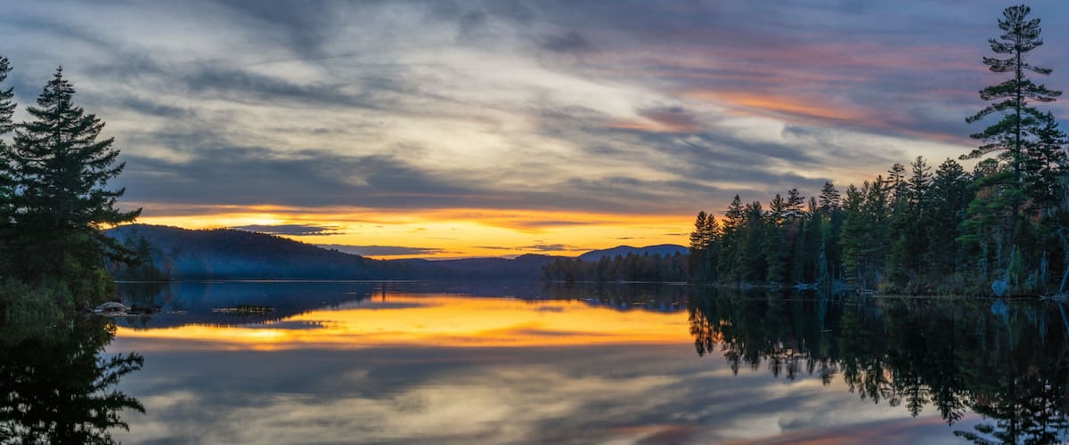 A serene lake at sunset with reflections of trees and mountains.
