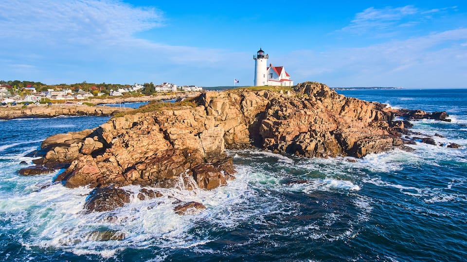 A lighthouse on a rocky outcrop with waves crashing against it, surrounded by a coastal town.