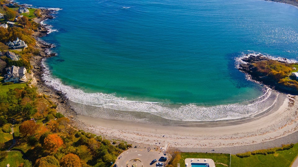 A coastal landscape with a beach, a parking area, and a swimming pool.