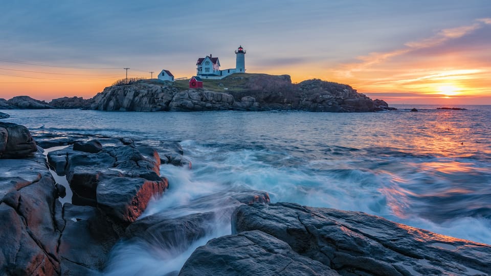 A coastal scene with a lighthouse, a house, and rocky shores at sunset.