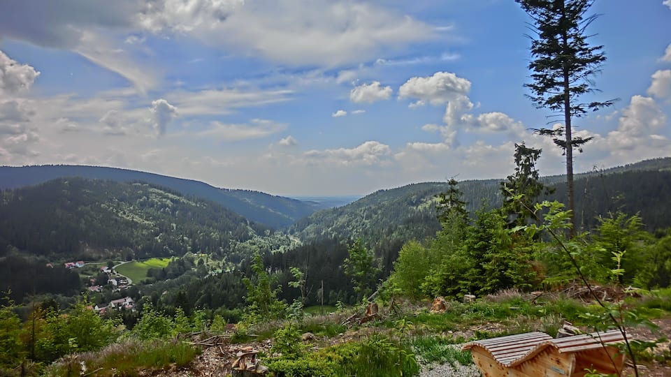 A valley with a wooden structure, dense forest, and distant hills under a partly cloudy sky.