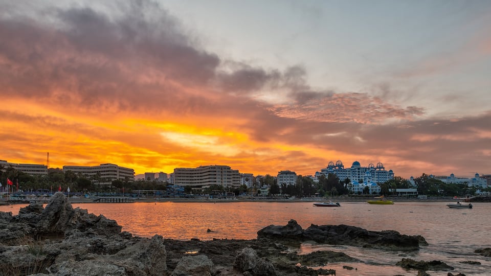 A sunset over a coastal city with rocky shores, boats, and a large hotel with domed architecture.