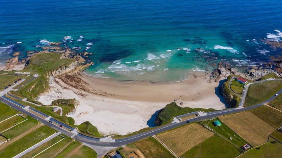 An aerial view of a coastal area with a sandy beach, rocky outcrops, and a road leading to a small settlement.