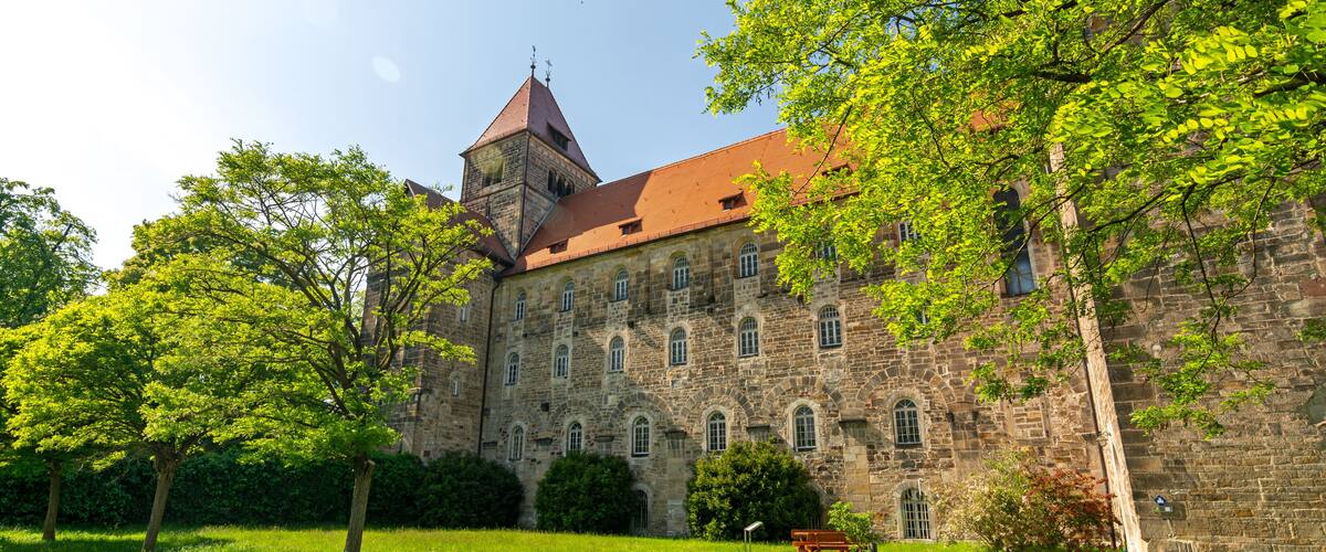 A historic stone building with a red roof, surrounded by lush green grass and trees.