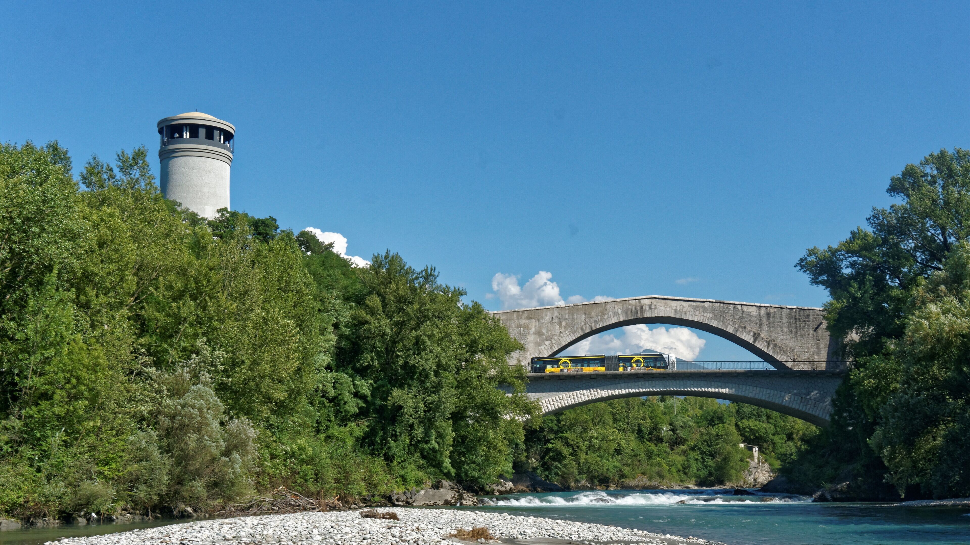 A stone bridge over a river, a lighthouse, and dense green trees.