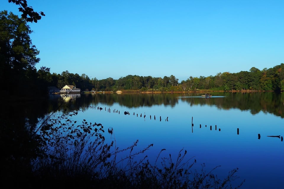 Un lago tranquilo con un pequeño muelle y un pabellón a la izquierda, rodeado de árboles frondosos y cielo azul despejado.