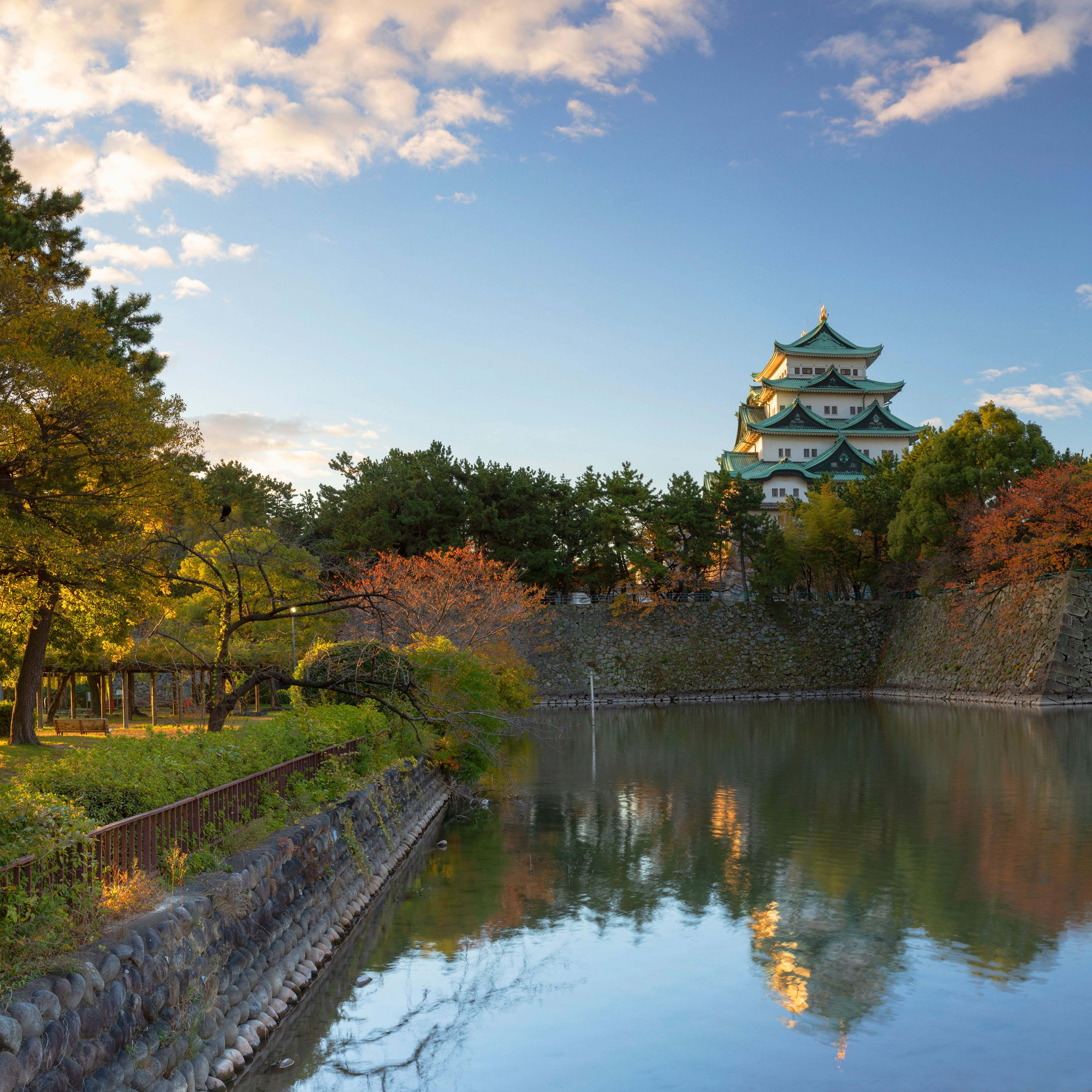 Eine traditionelle japanische Burg mit mehreren Ebenen, umgeben von Herbstlaub und einem stillen See, der den Himmel spiegelt.