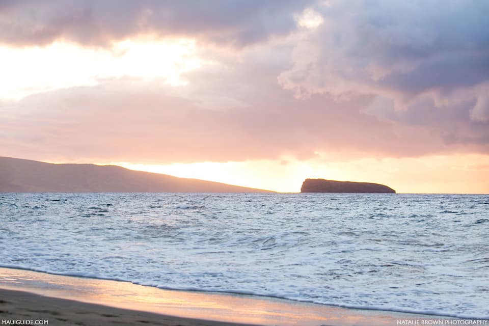 A beach with waves and a distant rocky outcrop during sunset.