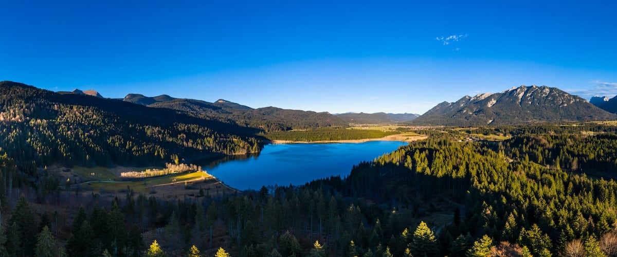 A mountainous landscape with a lake surrounded by dense forests and distant peaks.