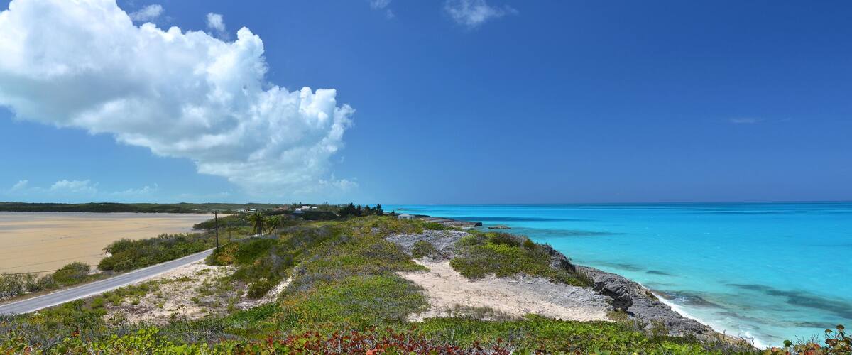 A coastal landscape with a road leading to a beach, a large cloud in the sky, and a clear blue ocean.
