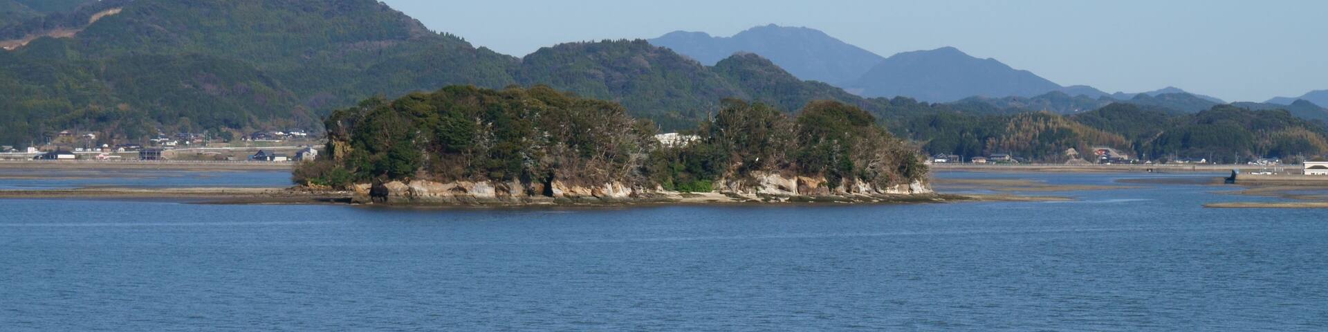 A calm lake with a small island covered in trees, surrounded by mountains and a distant shoreline with buildings.