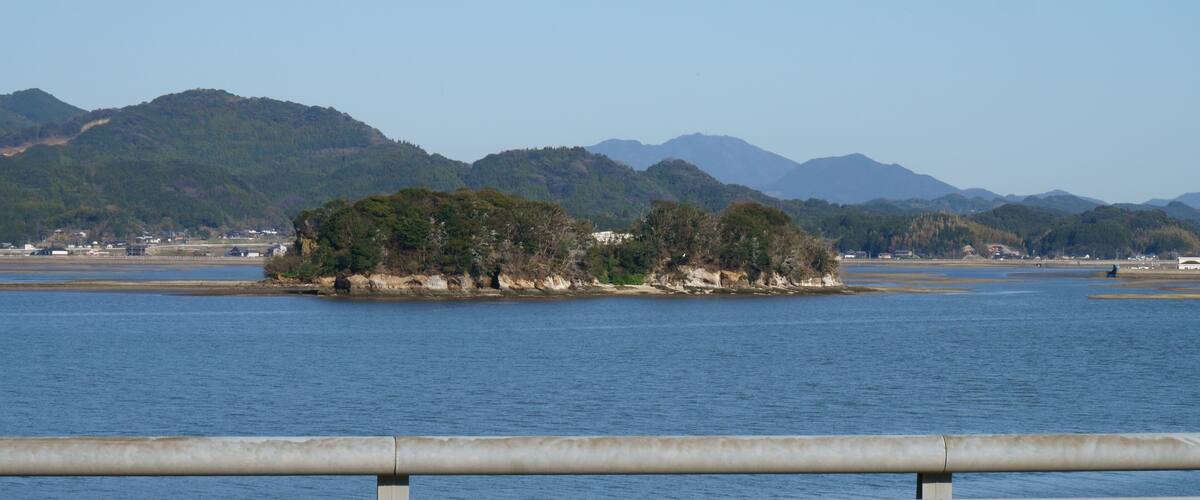 A calm lake with a small island covered in trees, surrounded by mountains and a distant shoreline with buildings.