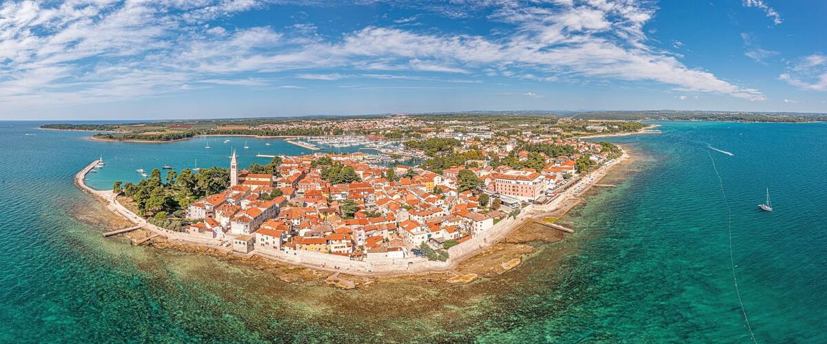 An aerial view of a coastal town with a clear blue sea, a prominent church tower, and a well-preserved harbor with boats.