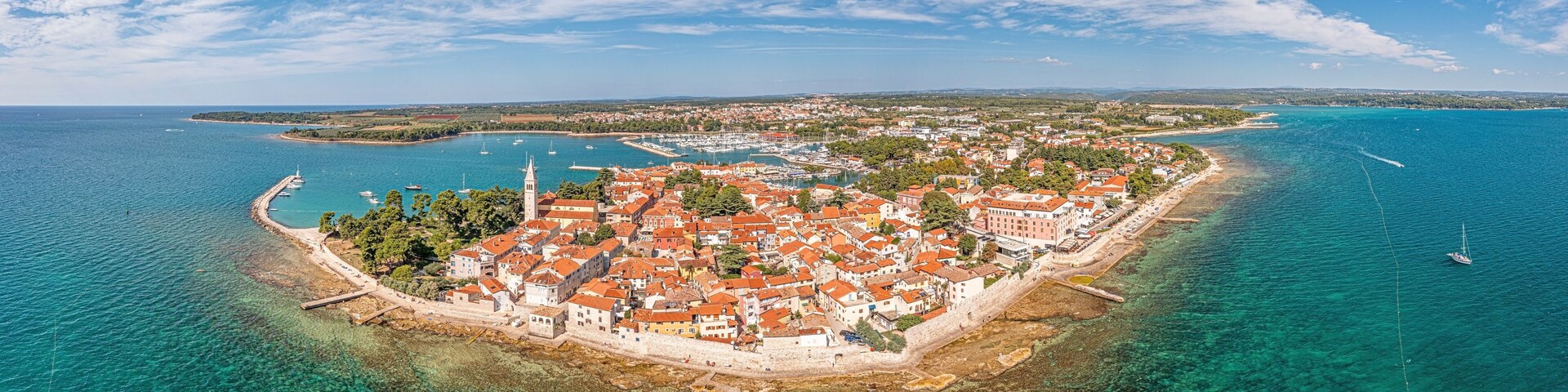 An aerial view of a coastal town with a clear blue sea, a prominent church tower, and a well-preserved harbor with boats.