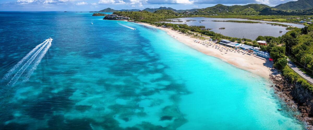A turquoise beach with clear waters, a boat creating a wake, and a backdrop of green hills and a clear sky.