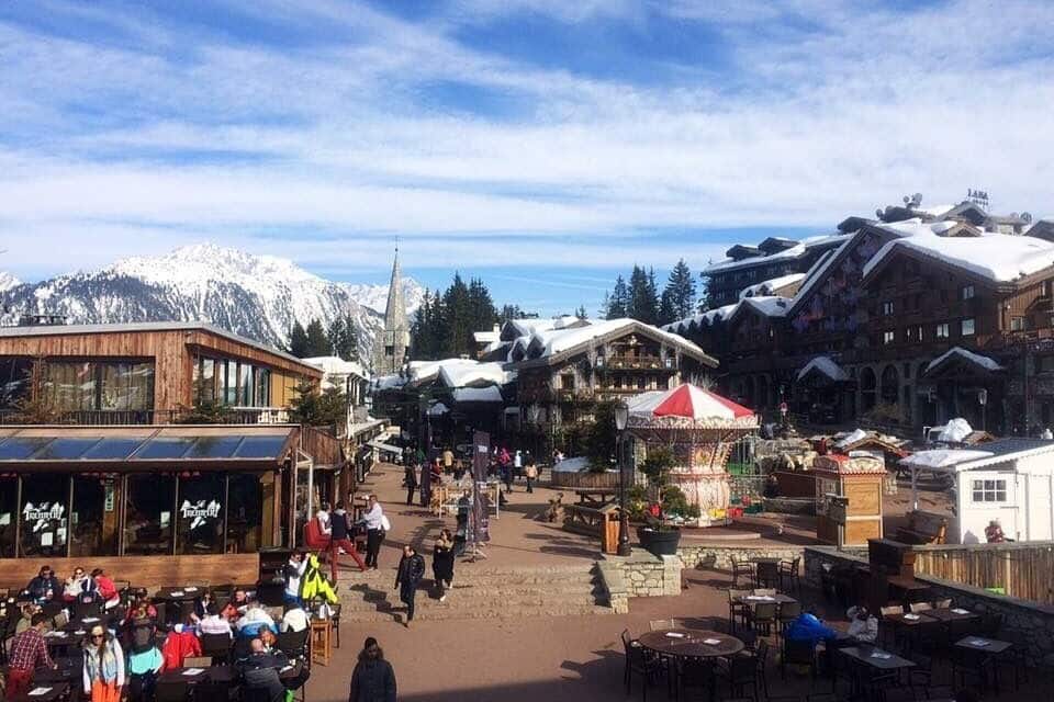 Un pueblo en la montaña cubierto de nieve con edificios de madera, mesas al aire libre y cielo despejado.