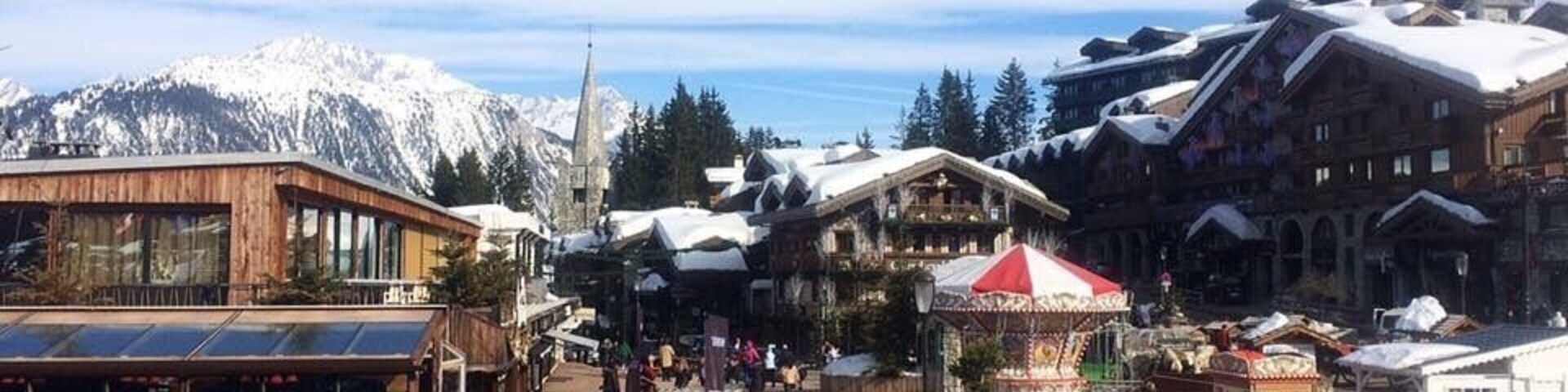 Un pueblo en la montaña cubierto de nieve con edificios de madera, mesas al aire libre y cielo despejado.