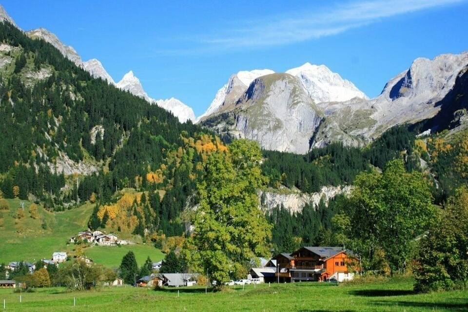 A mountainous landscape with a village nestled among green fields and trees, with snow-capped peaks in the background.