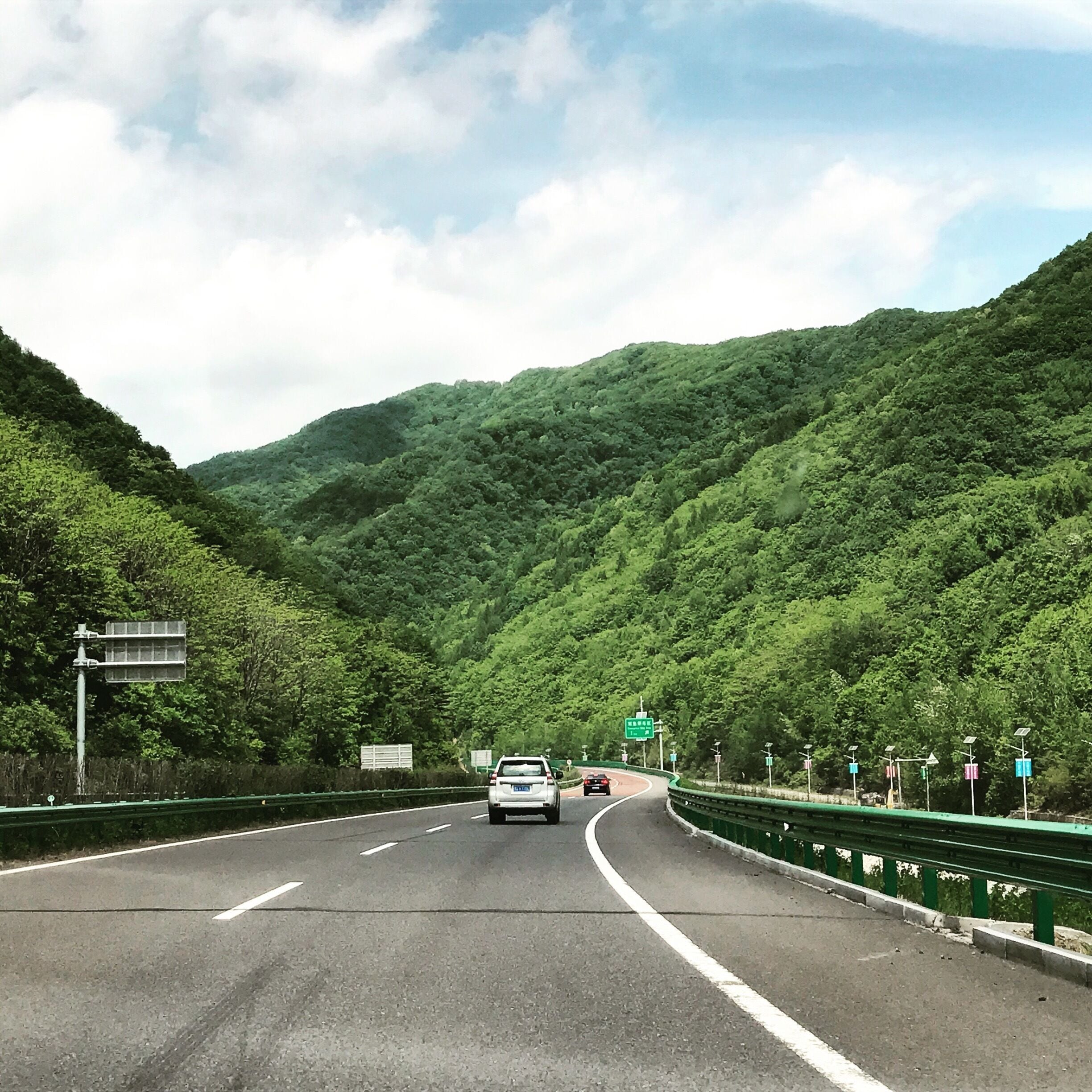 A highway with green guardrails, surrounded by lush green hills.