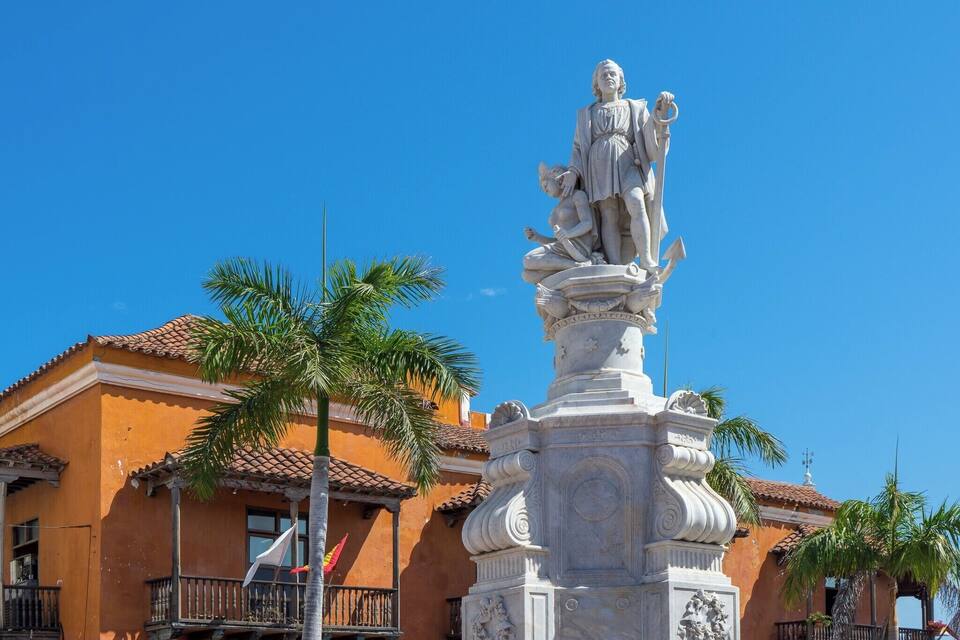 A statue of a historical figure with a child, set against a building with a tiled roof and palm trees.