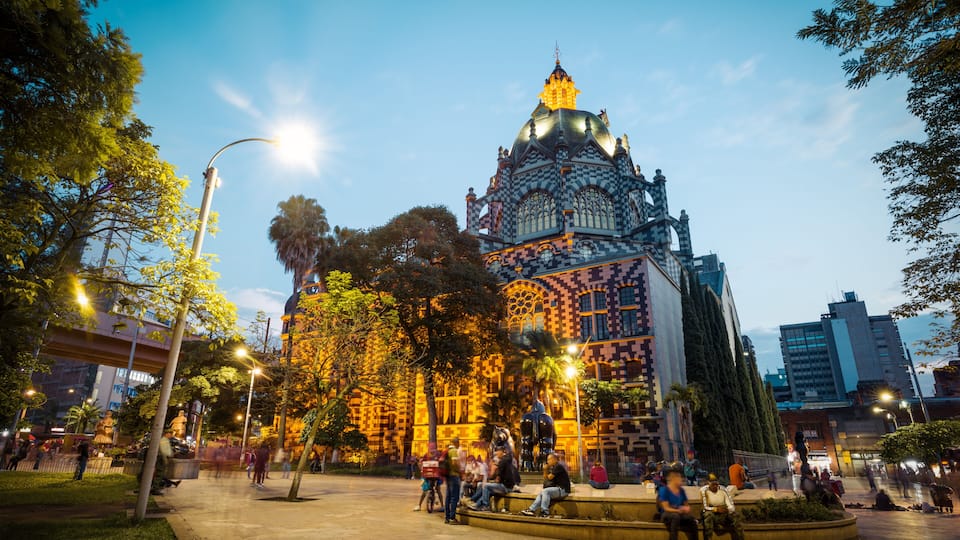 A historic building with intricate architecture and a large dome, surrounded by a plaza with people and trees.