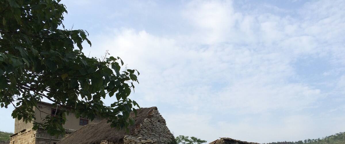A stone house with a thatched roof, surrounded by greenery and hills in the background.