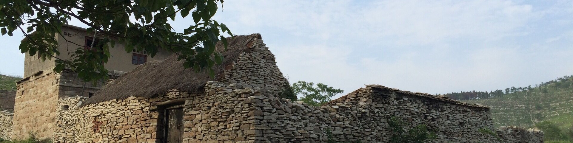 A stone house with a thatched roof, surrounded by greenery and hills in the background.