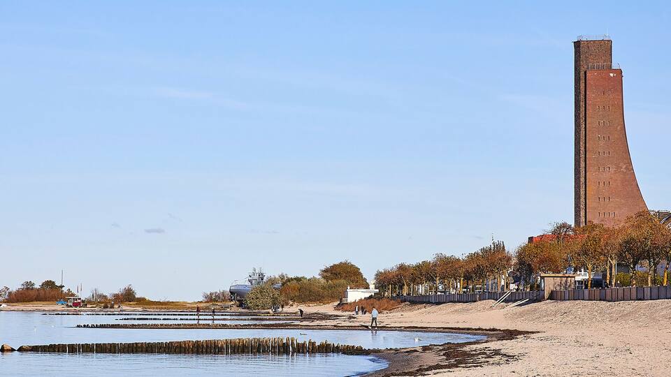 A coastal area with a tall brick tower, sandy beach, and autumn trees.