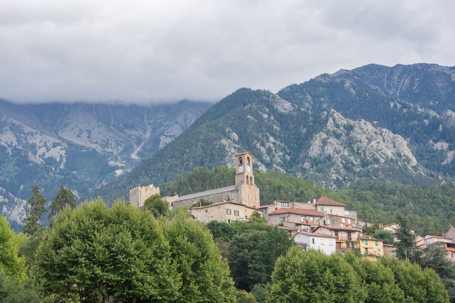 Un paisaje montañoso con una iglesia histórica que cuenta con una torre del reloj, rodeado de exuberante vegetación y edificios residenciales.