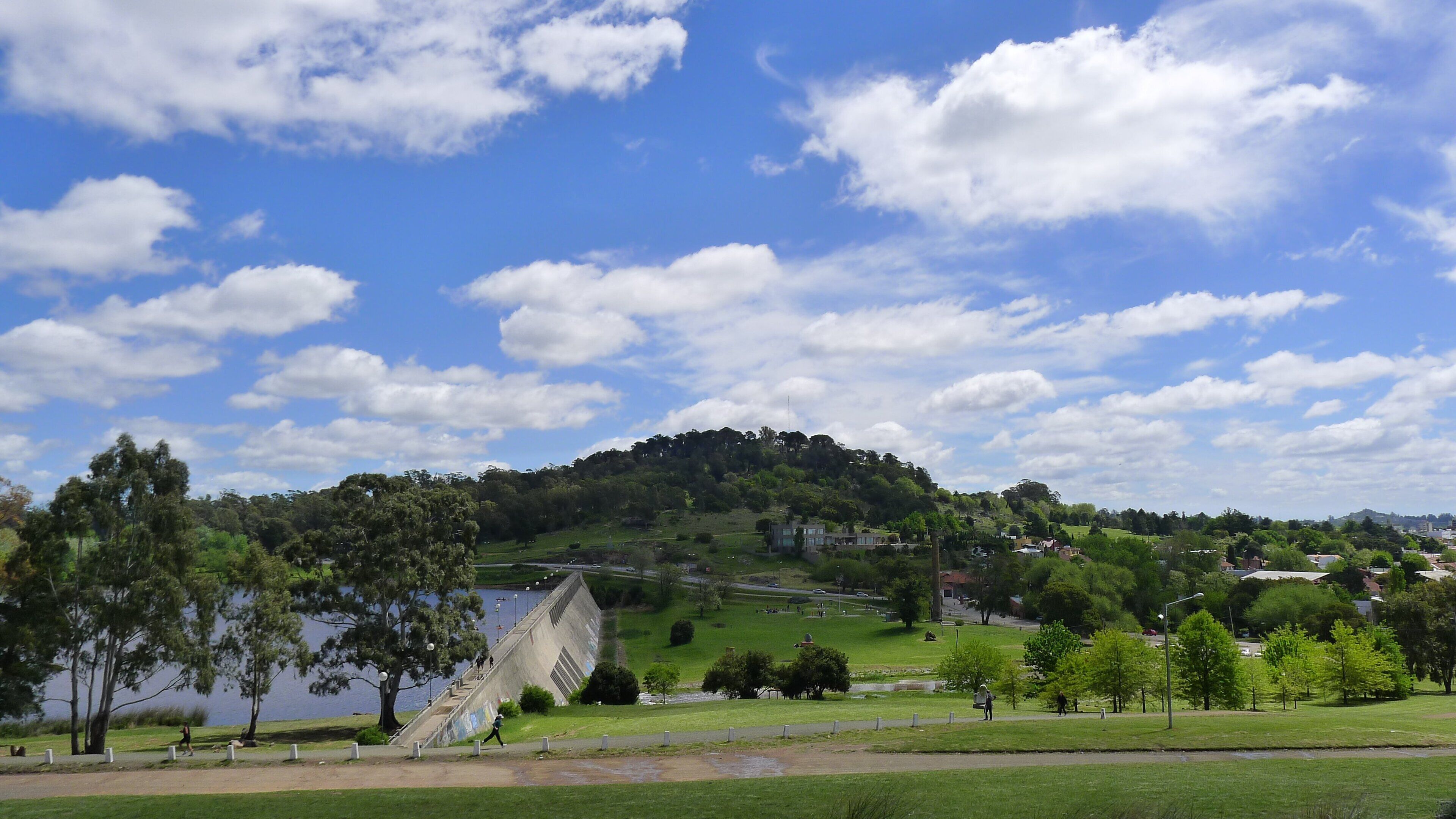 A park with a lake, a concrete structure, and a hill with trees.