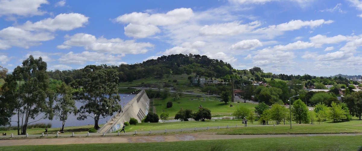 A park with a lake, a concrete structure, and a hill with trees.
