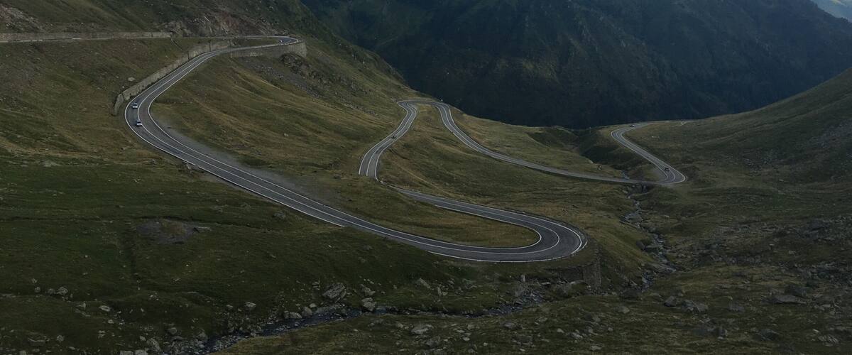 A winding mountain road surrounded by lush green hills and towering mountains under a clear blue sky.