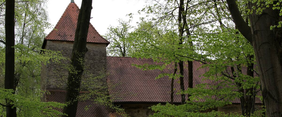 A medieval stone building with a red-tiled roof, surrounded by lush green trees.