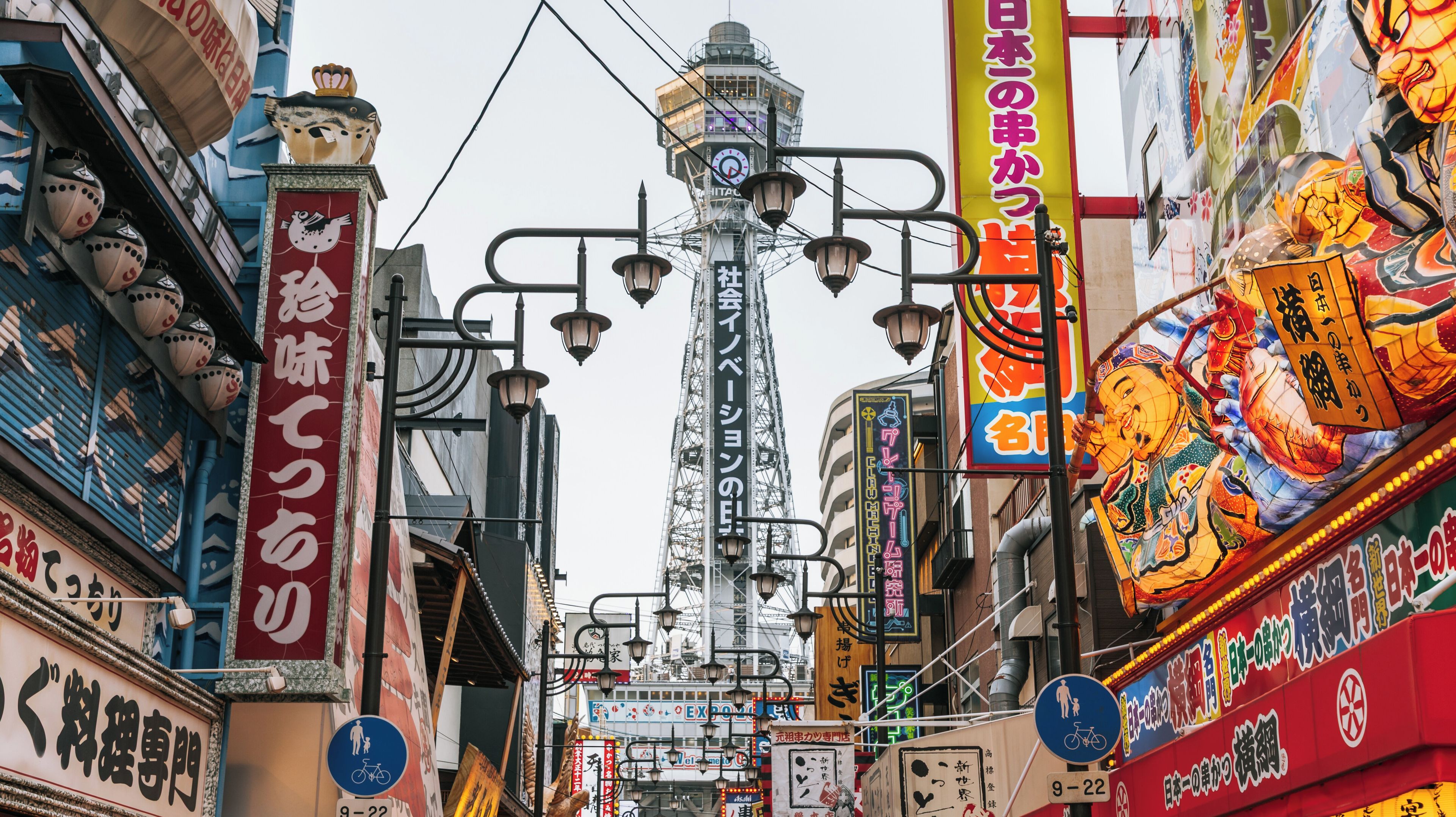 Uma rua urbana movimentada com uma torre imponente, sinalização colorida e tradicionais lanternas japonesas.